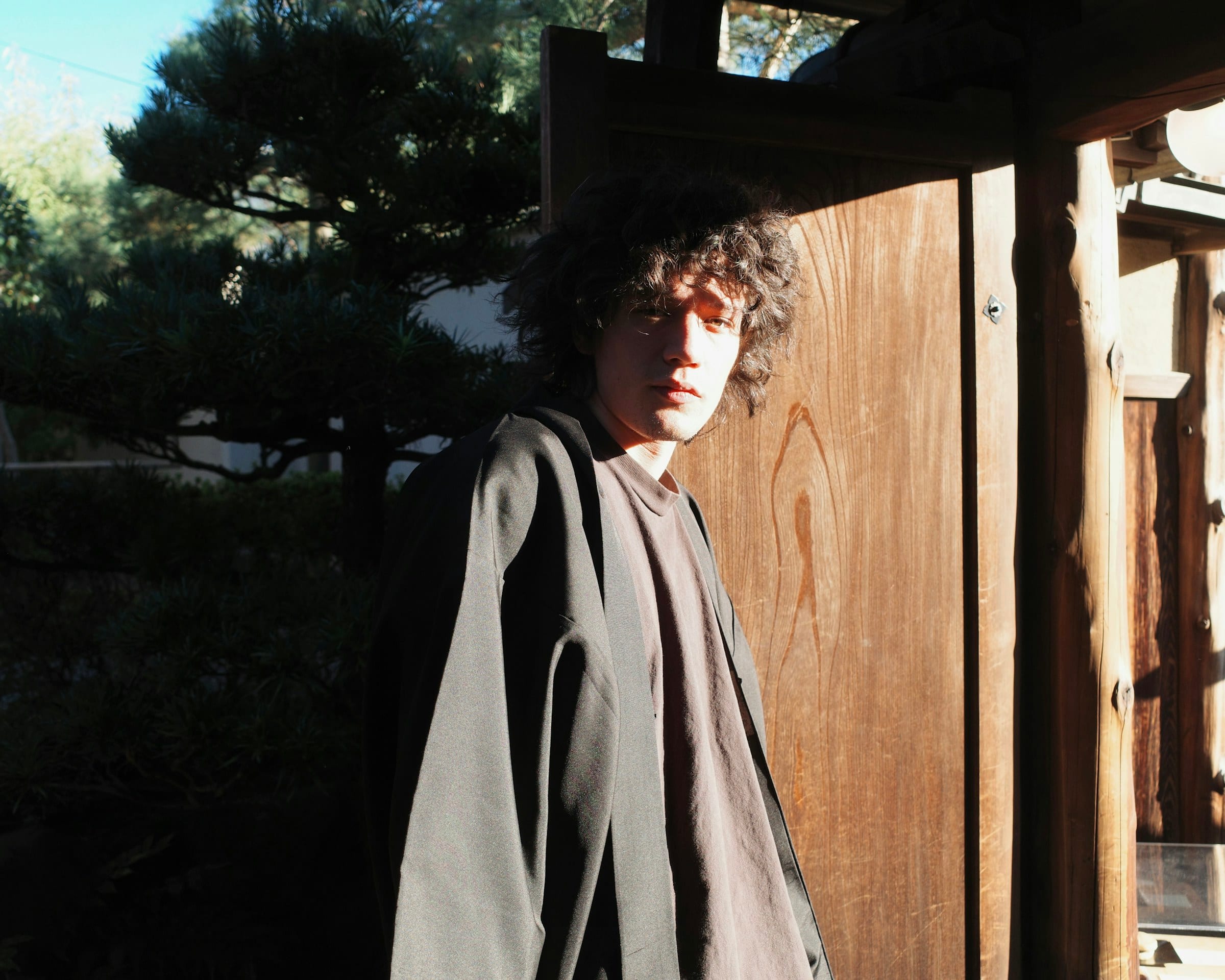 A young man in a grey coat standing in a sunlit wooden doorway in Kyoto