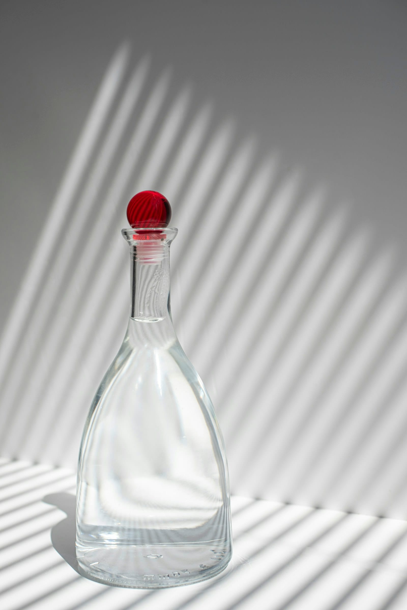 A glass decanter with a red stopper casting long striped shadows