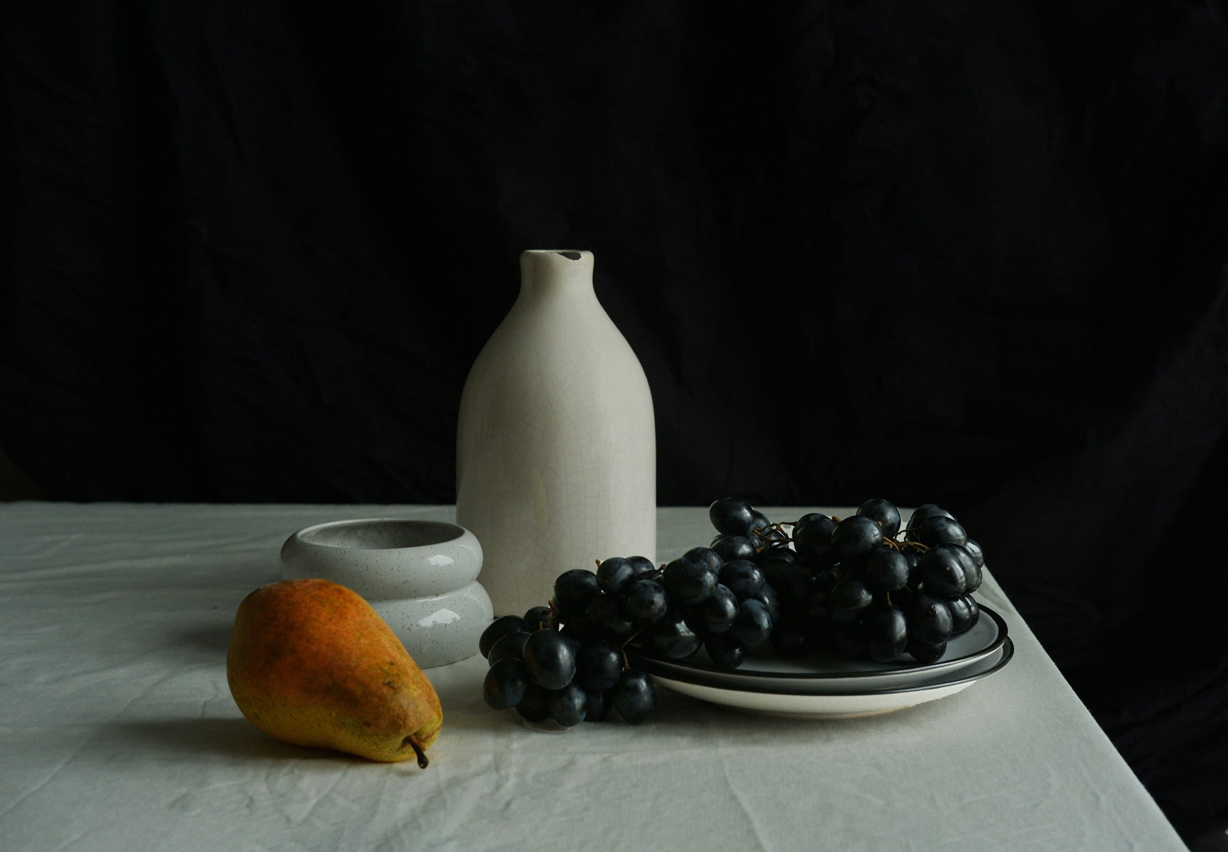 A still life of a white vase, a pear and grapes on linen against black